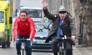 Chris Boardman cycling with Boris (in a very snazzy hat)