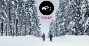 Two people cycling through snow, a bleak grey sky is behind them