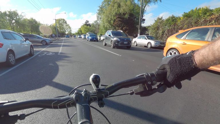 View from rider's handle bars of them taking the centre of the traffic lane, making them more visible to a car ahead who is waiting to turn right.