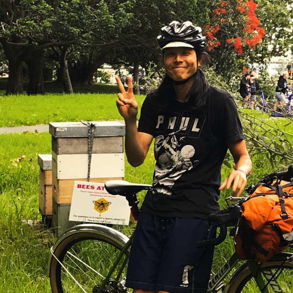 A photo of Teva Chonon with one hand up in a peace sign. They are leaning on a bicycle. There is grass, pohutakawa trees, and a community beehive in the background.