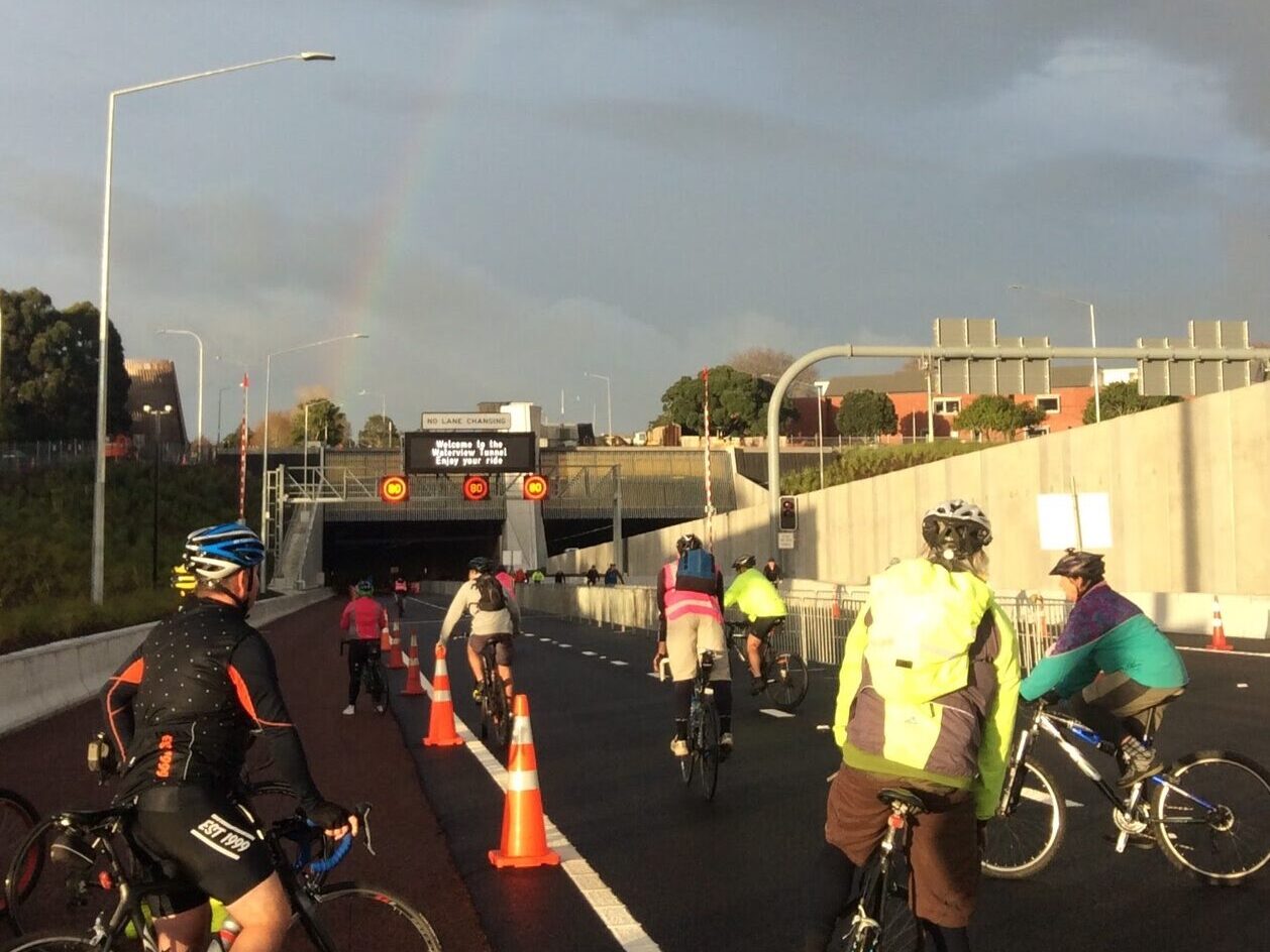 Many people using bikes to go through on the Waterview Tunnel. There is a rainbow in the sky overhead.