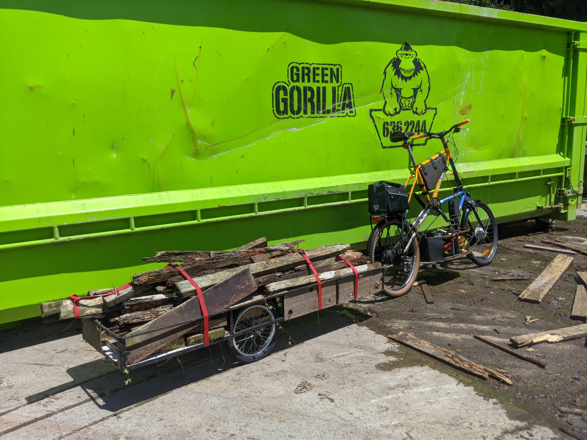A tall bike with bike trailer posed in front of a large green gorilla waste bin. The bike trailer is loaded with scrap planks of wood.