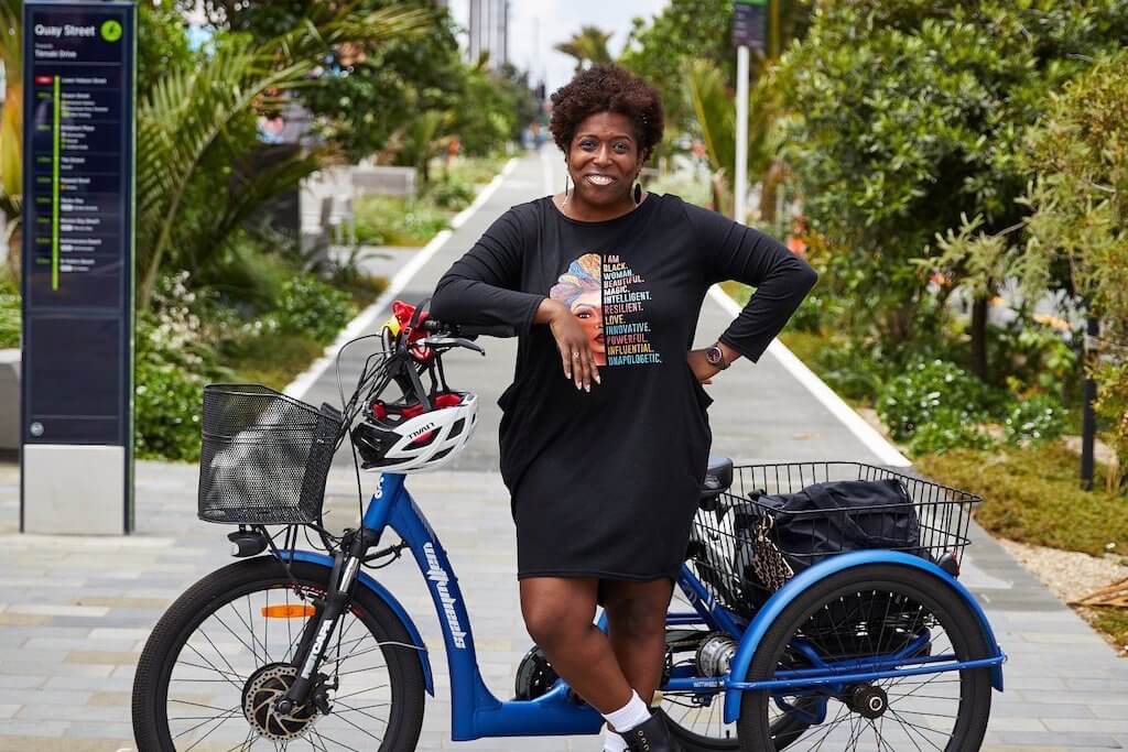A photo of Tiffany Robinson, leaning on he blue electric trike. The Quay Street cycleway in Auckland is in the background. 