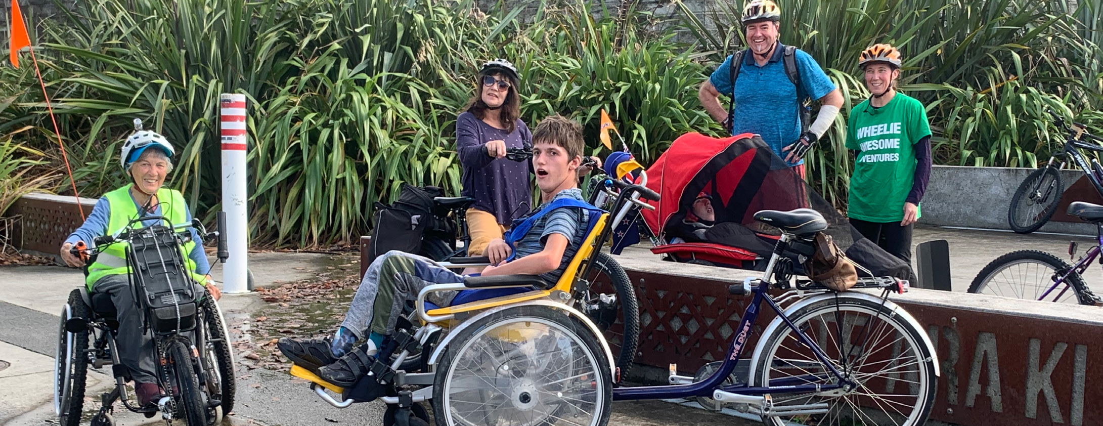 A group of five people. One is sitting in a wheelchair bike, another is using a wheelchair with power attachment, and the other three are standing behind them