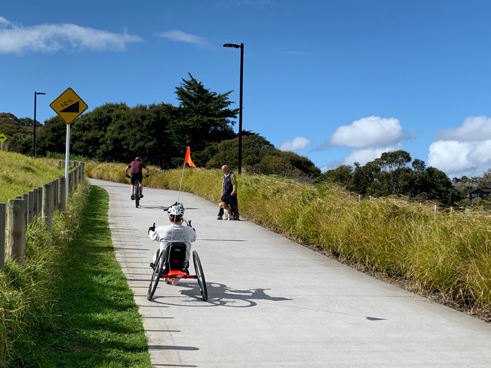 A person cycles up a hill using a recumbent handcycle. Ahead of them is someone cycling up the hill using a two-wheeled bike. It appears to be a section of Te Ara Ki Uta Ki Tai, the Glenn Innes to Tāmaki Pathway.