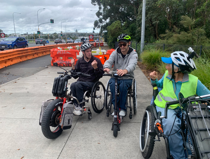 Three people using wheelchairs with powered attachments are chatting with each other. In the background is a detour. It is recognisable as the section of Great North Road that connects to the Waterview pathway.