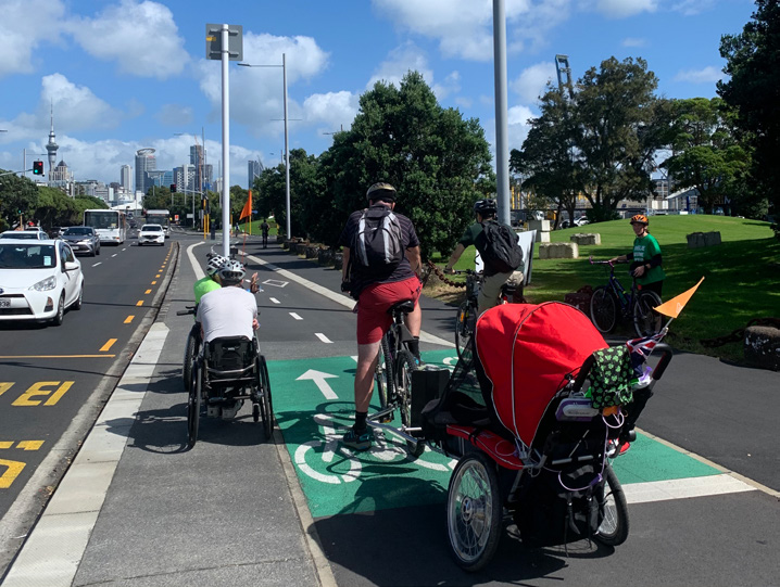 A group of people on bikes on the Tāmaki Drive protected cycleway. One person is on a two wheeled bike with a bike trailer attached, another couple are using wheelchairs with powered attachments. 
