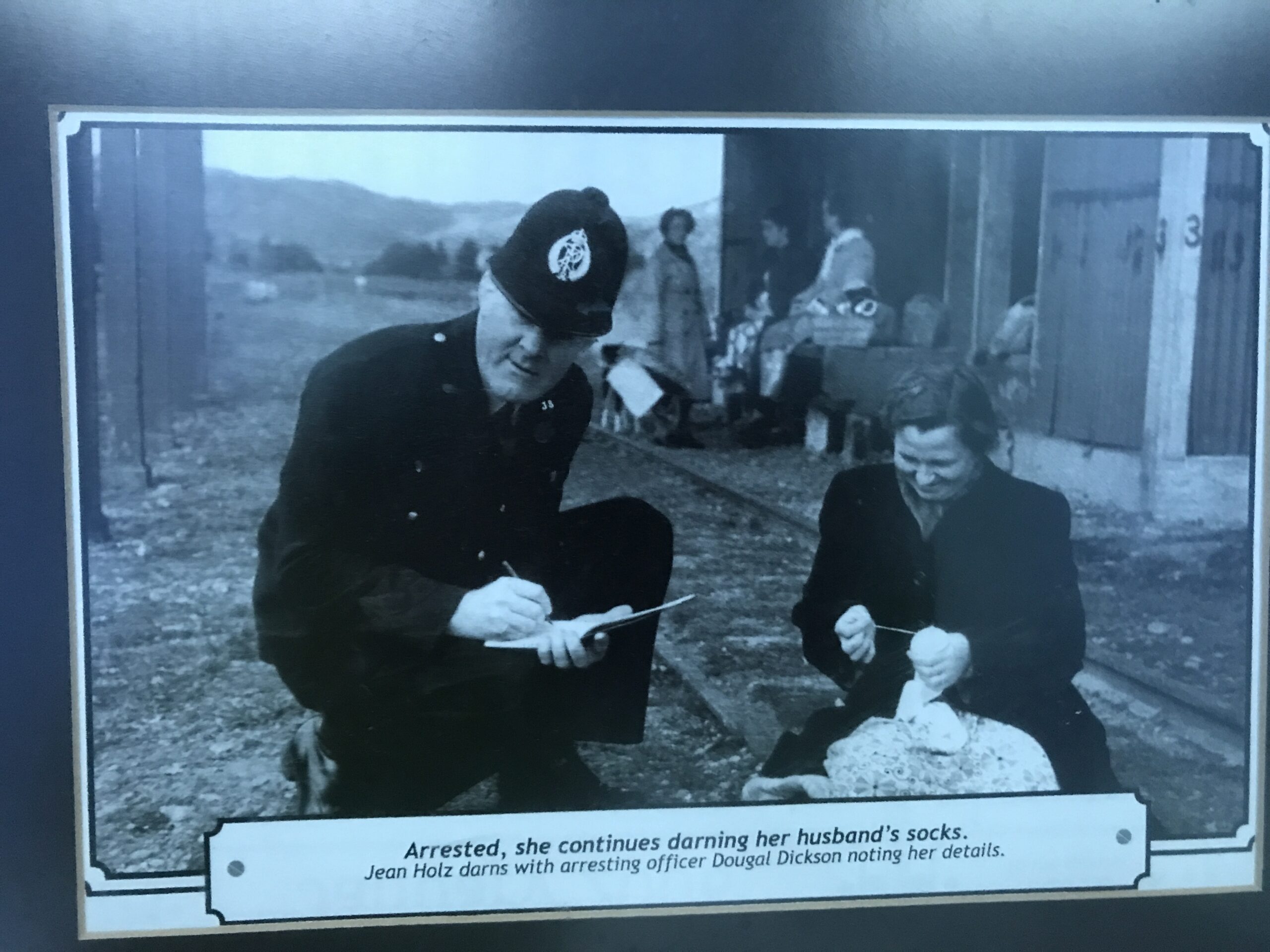 A lady sits on some railroad tracks darning socks. She's looking downwards and smiling. A police officer crouches beside her and looks at the camera while writing something in a booklet. The caption at the bottom reads "arrested, she continues darning her husband's socks. Jean Holz darns with arresting officer Dougal Dickson noting her details"