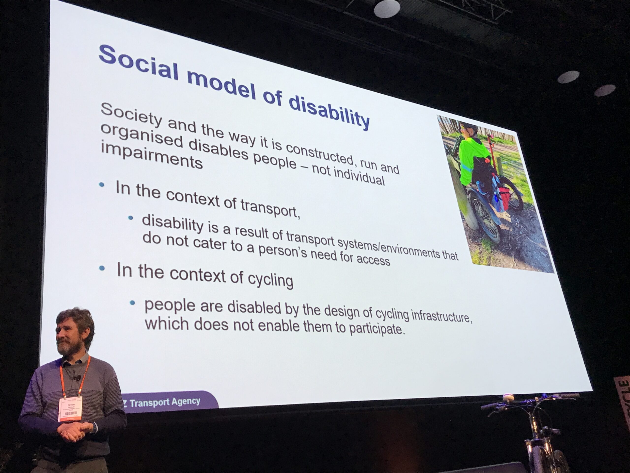 The slide behind Simon Kennett says "Social model of disability. Society and the way it is constructed, run and organised disables people - not individual impairments. In the context of transport disability is a result of transport systems/environments that do not cater to a person's need for access. In the context of cycling people are disabled by the design of cycling infrastructure which does not enable them to participate. 