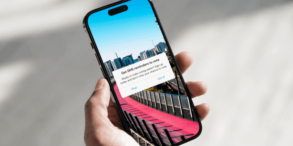 A hand holds a phone with a popup saying "get SMS reminders to vote"