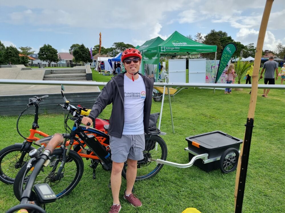 Bobby smiles in front of a bike with a trailer. In the background are some event marquees