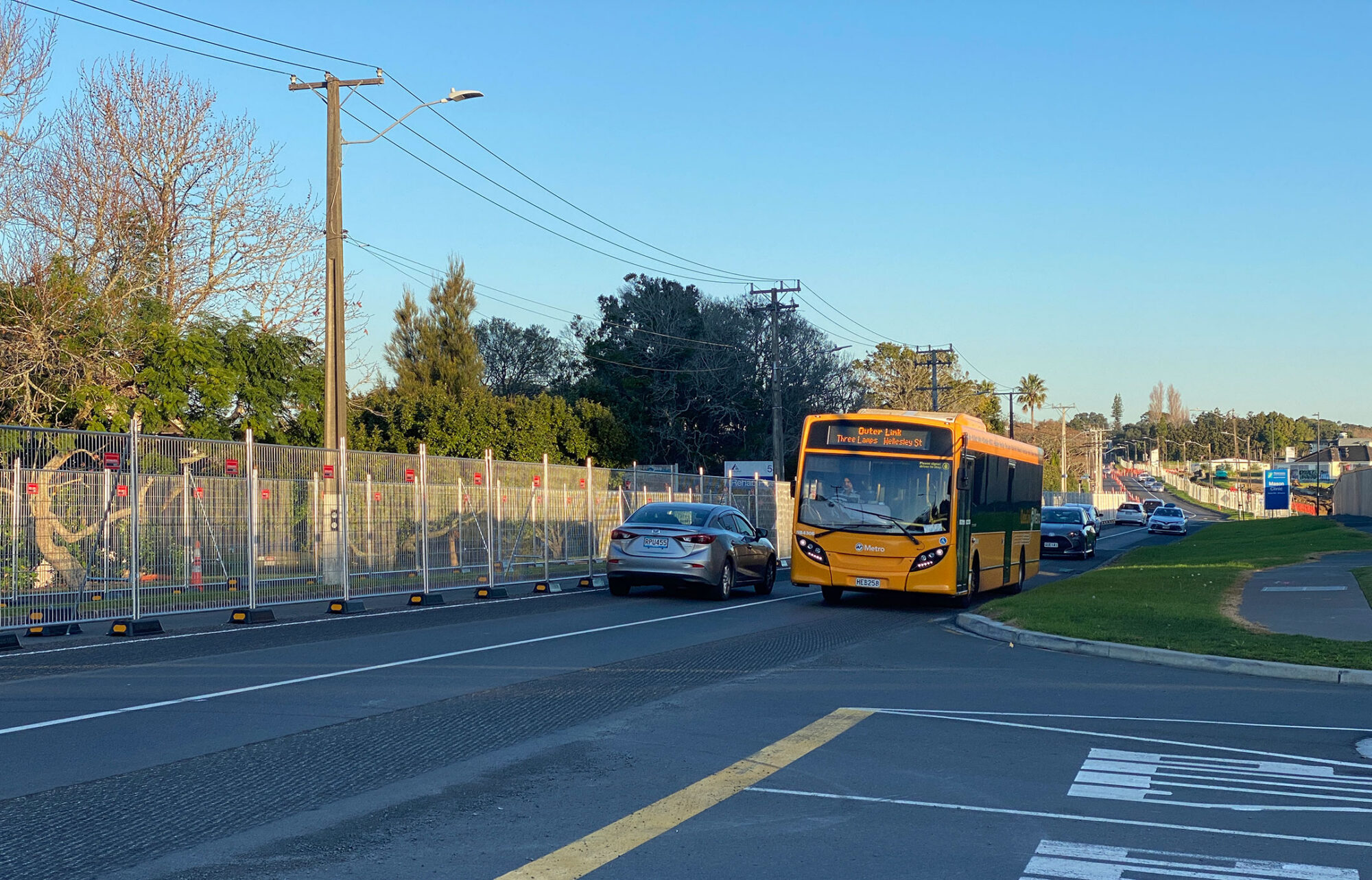 A road with a yellow bus coming towards the photographer. A car travels in the opposite direction. There's a construction fence along the side of the road and the road surface is very bumpy